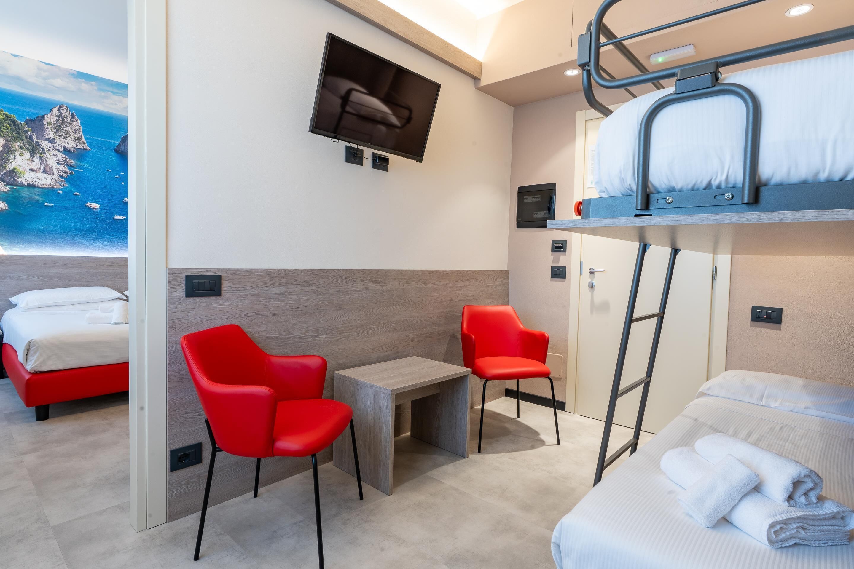 Une chambre d’hôtel compacte avec un lit superposé, deux lits simples, une petite table, deux chaises rouges, un téléviseur fixé au mur et une vue magnifique sur l’océan.