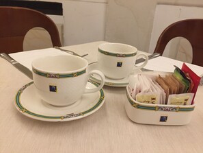 Una mesa servida con dos tazas blancas con detalles dorados y verdes, un platillo a juego y una pequeña cesta con bolsitas de té.