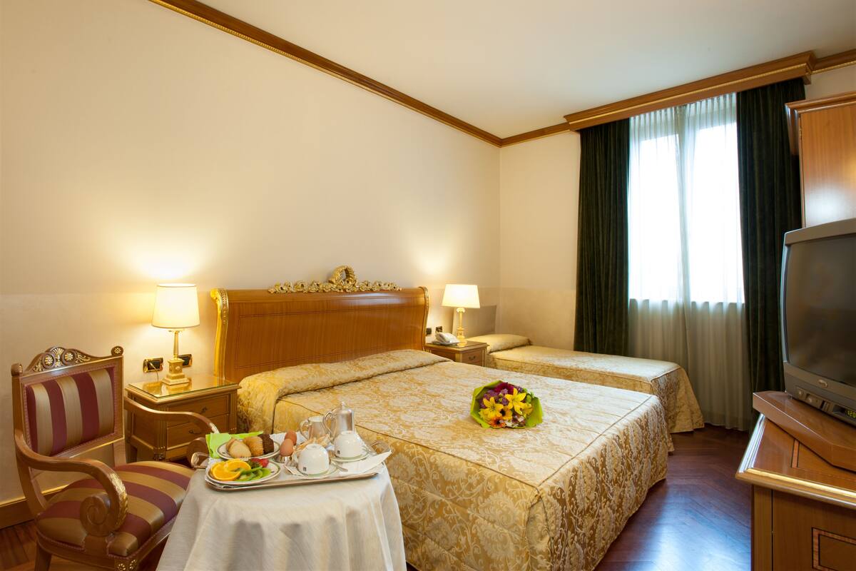 A hotel room with two beds, a television, a dining table with breakfast items, and a decorative chair.