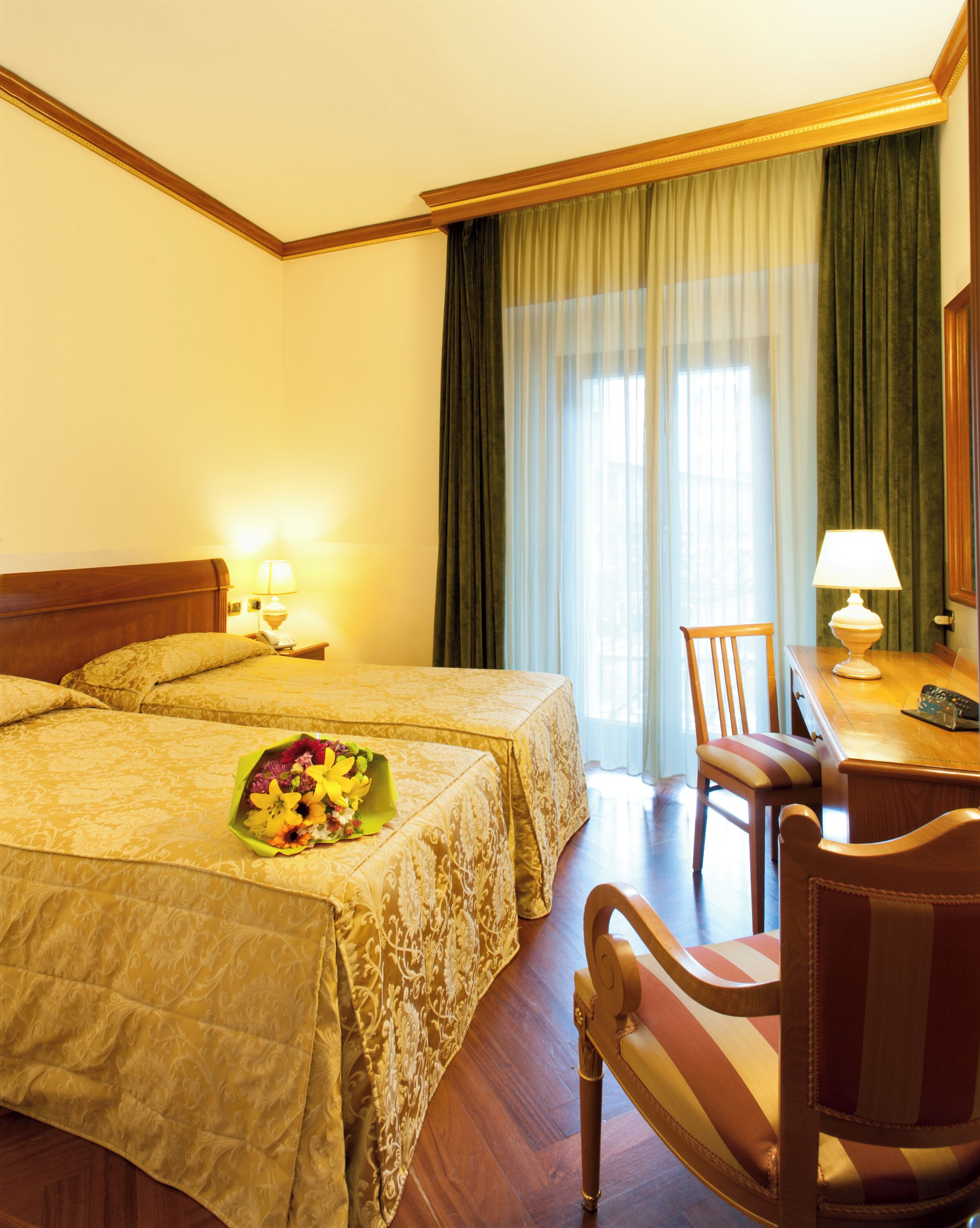 Habitación de hotel con dos camas, un escritorio con lámpara, una silla y un ramo de flores.