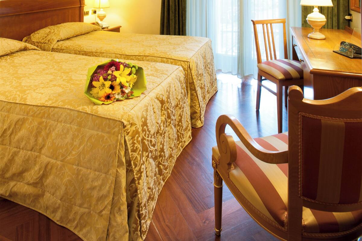 Habitación de hotel con dos camas, un escritorio con lámpara, una silla y un ramo de flores.
