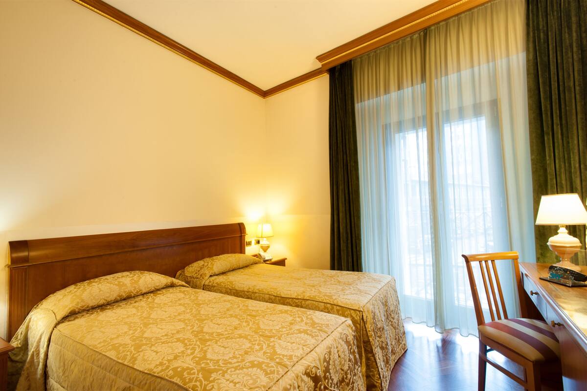A hotel room with two beds, a desk, a chair, a lamp, and a window with curtains.