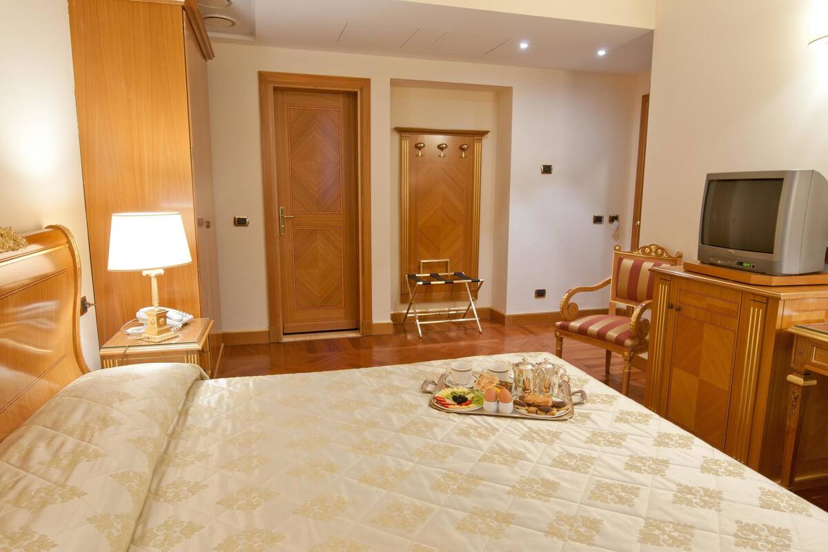 A hotel room with a bed, a bedside table with a lamp, a television, a chair, and a tray with breakfast items.