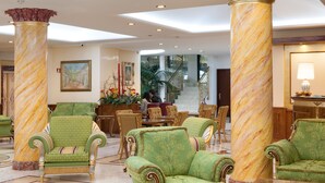 A lobby with green upholstered chairs, marble columns, and a staircase in the background.