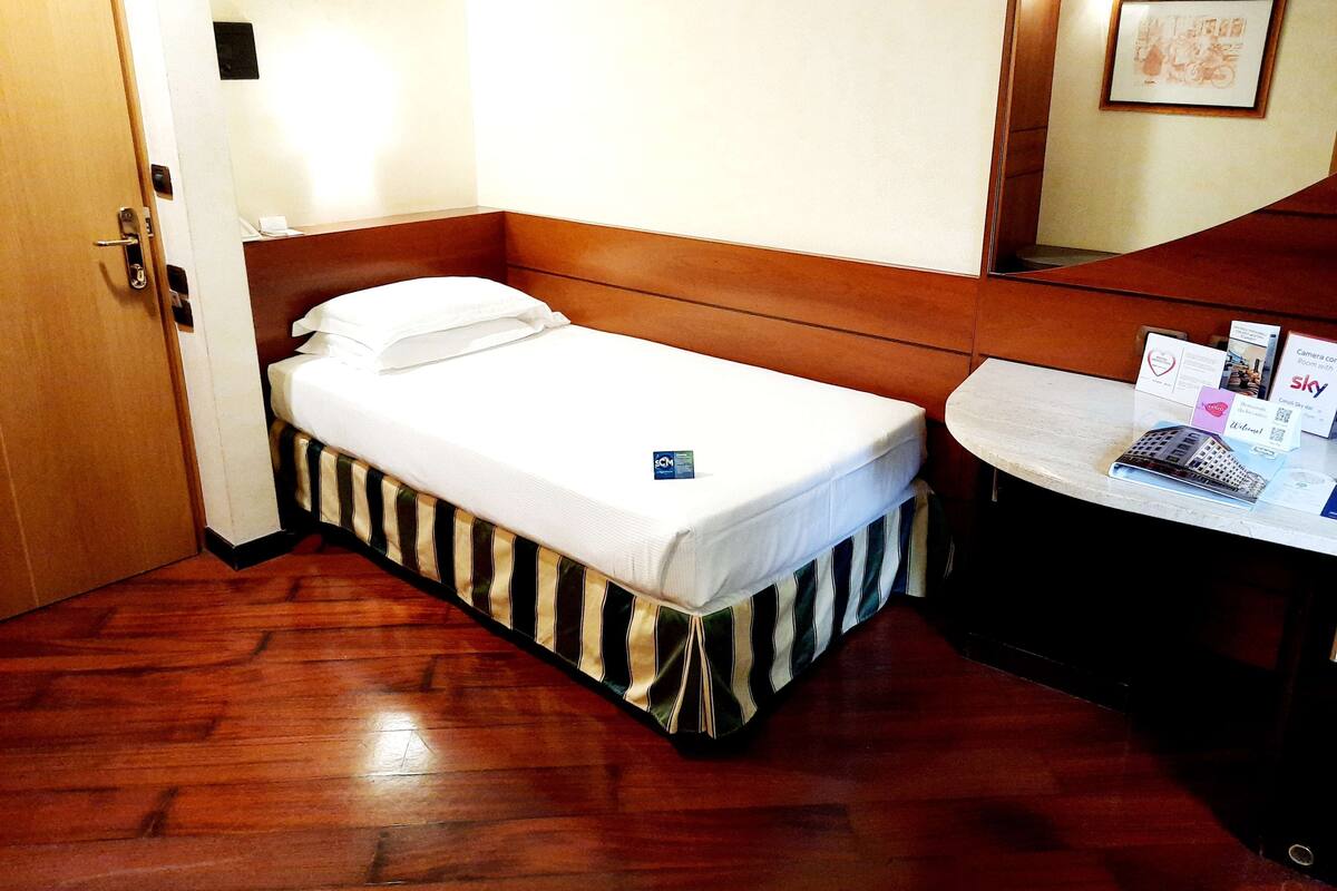 Een kleine hotelkamer met een enkel bed, een houten hoofdbord, een nachtkastje met een lamp en een spiegel.