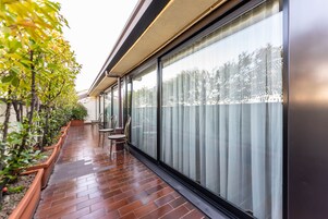 Un balcone con pavimento in mattoni, piante in vaso e una porta vetrata che dà su una vista alberata.