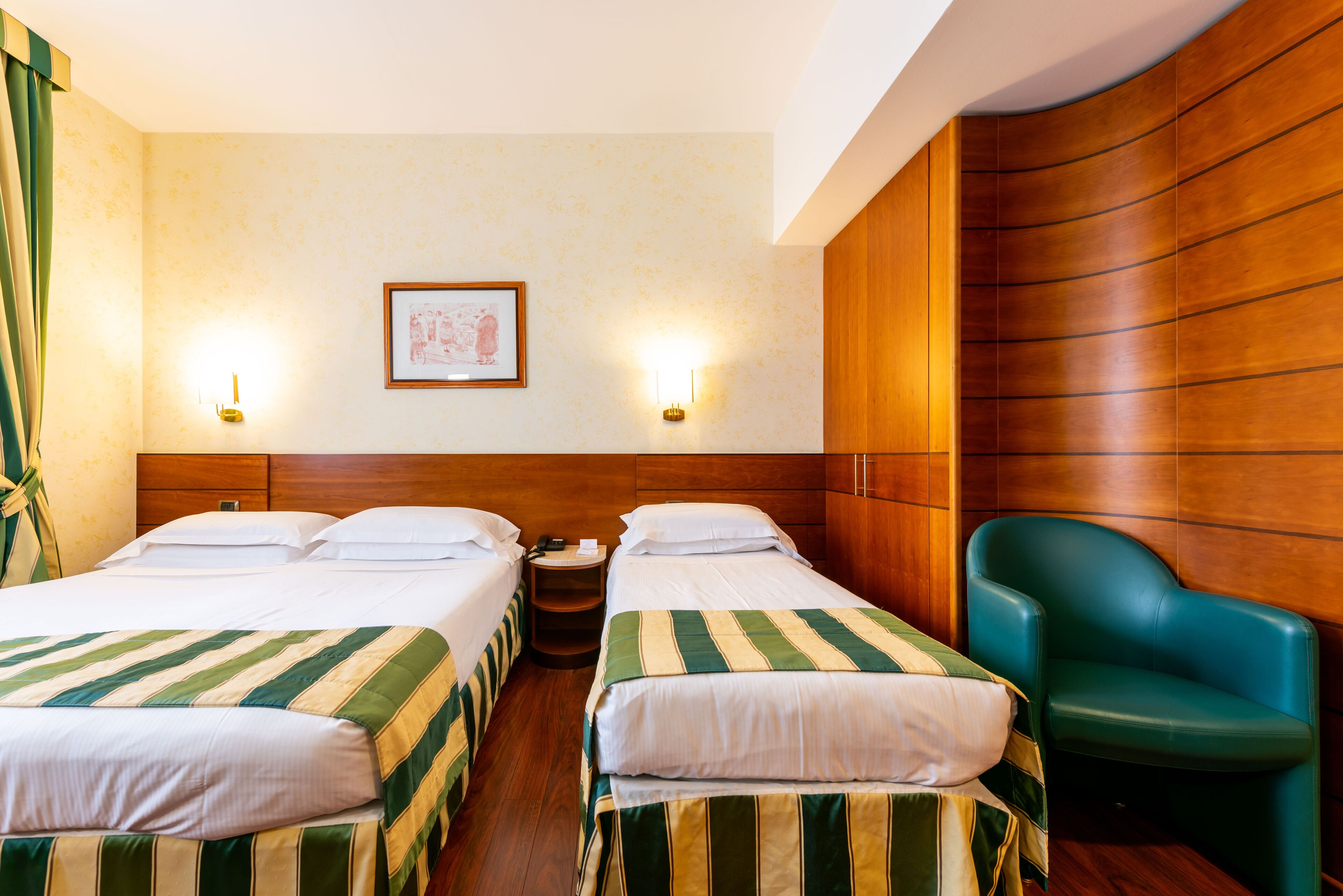 A hotel room with two beds, a wooden headboard, a green chair, and a framed picture on the wall.
