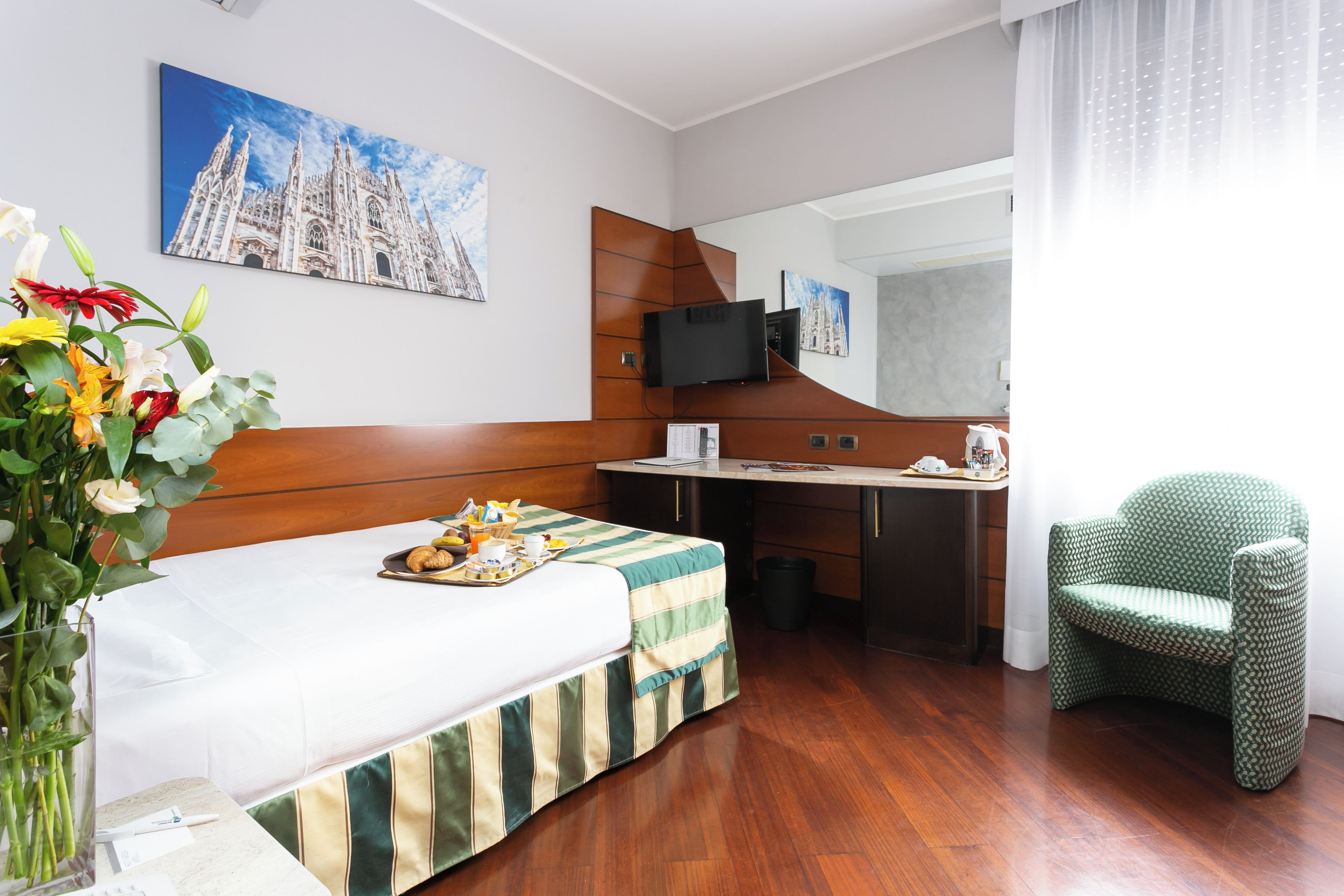 A hotel room with a bed, a desk, a chair, a TV, and a painting of a cathedral.