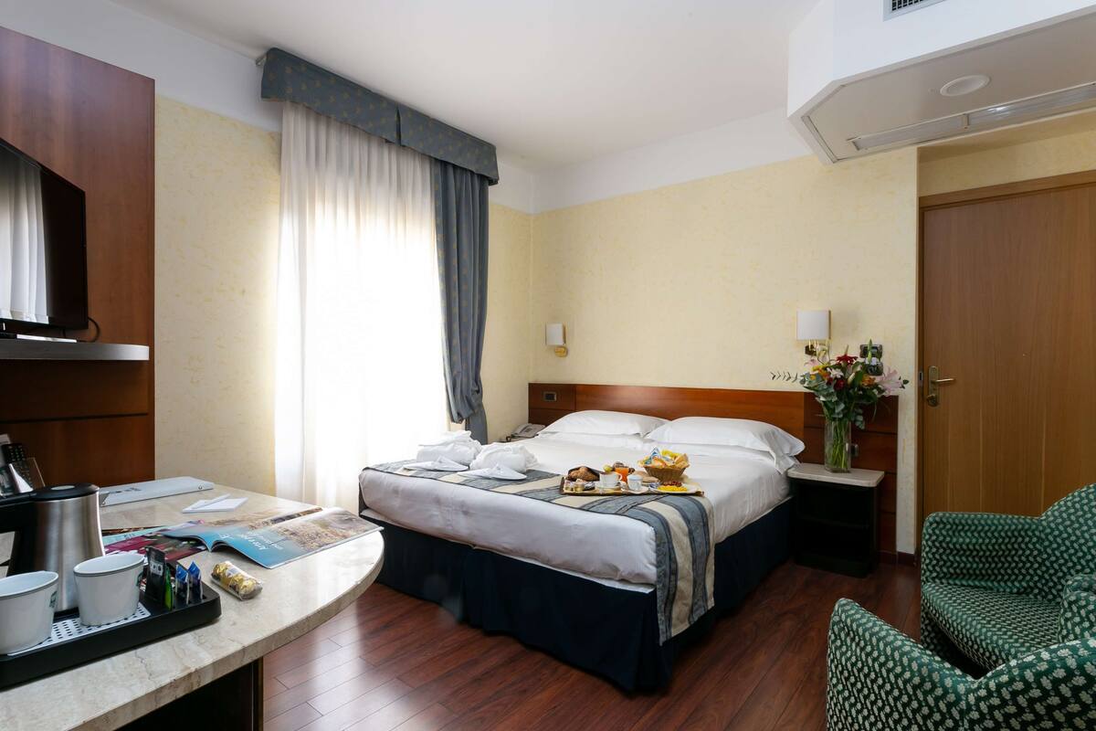 Een hotelkamer met een bed, een bureau met koffiezetapparaat, een stoel en een nachtkastje met lamp.