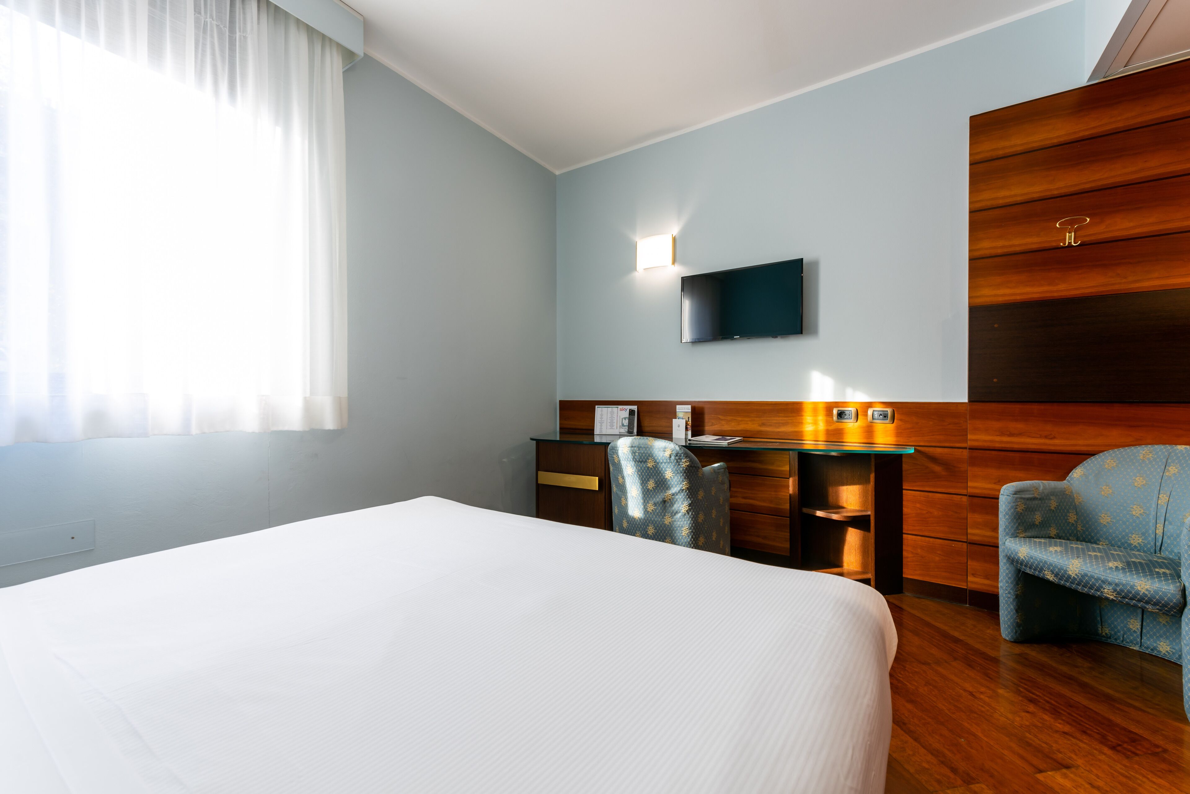 A hotel room with a large bed, a desk with a chair, a TV mounted on the wall, and a wooden panel on the right side.
