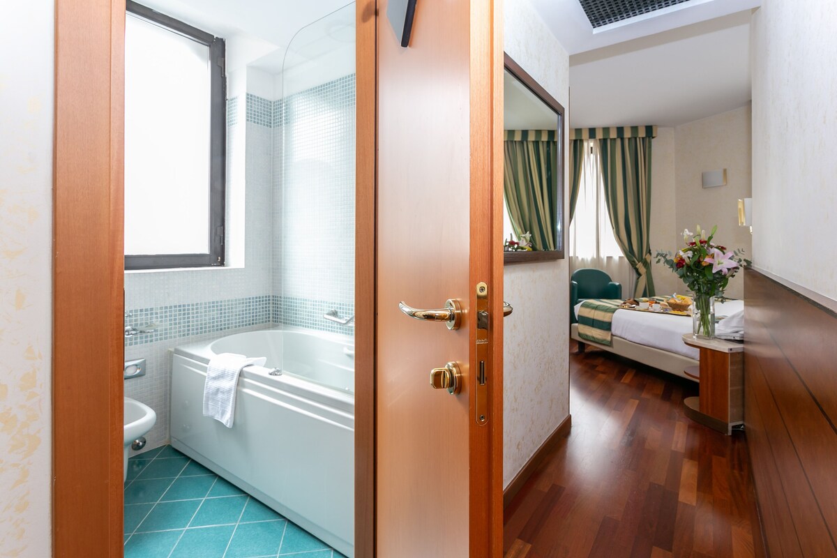 Een hotelbadkamer met een badkuip, wastafel en toilet. Een houten deur leidt naar een slaapkamer met een bed, nachtkastje en een vaas met bloemen.