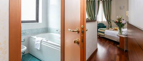 A hotel bathroom with a bathtub, sink, and toilet. There is a wooden door leading to a bedroom with a bed, nightstand, and a vase with flowers.