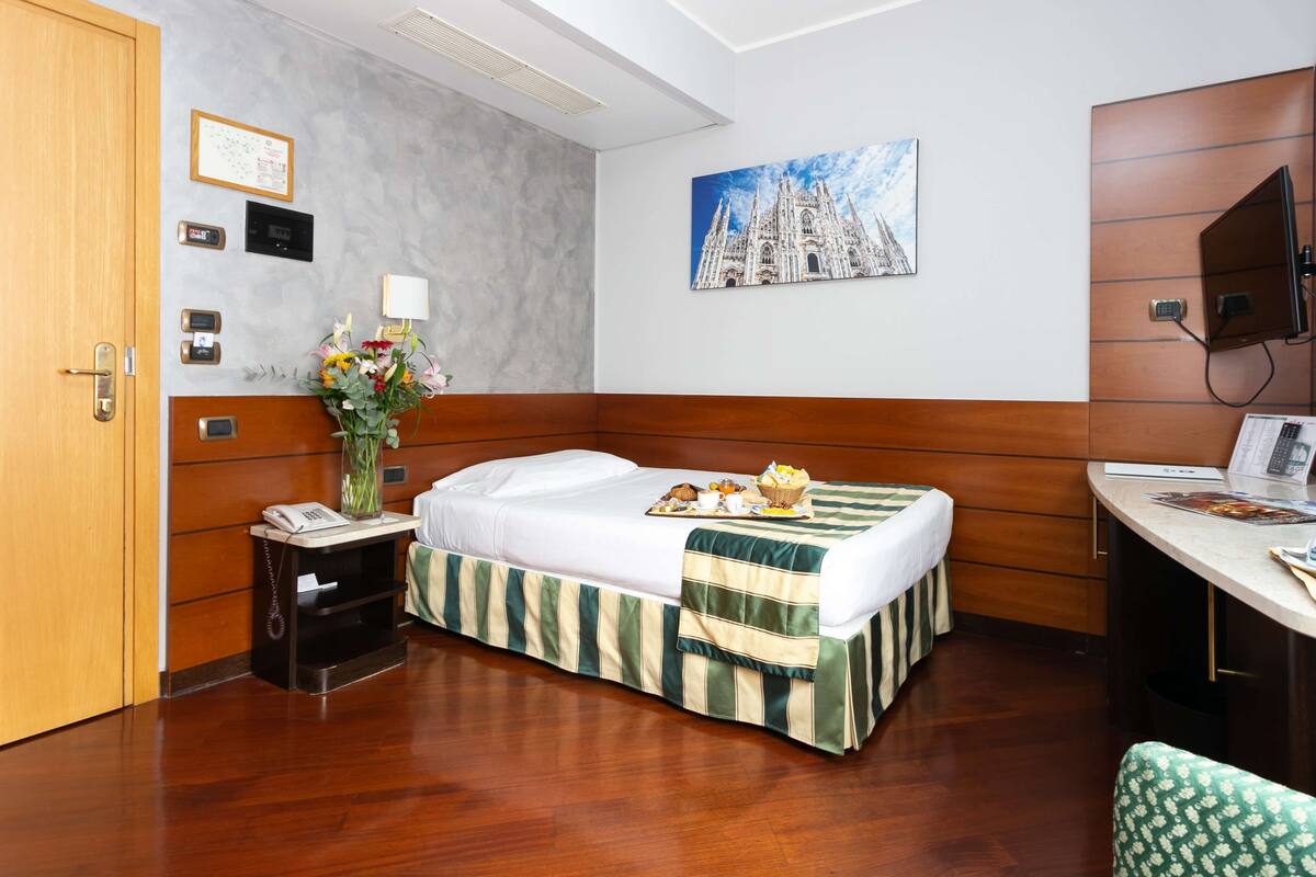 Een hotelkamer met een bed, nachtkastje, bureau en een schilderij van een kathedraal aan de muur.