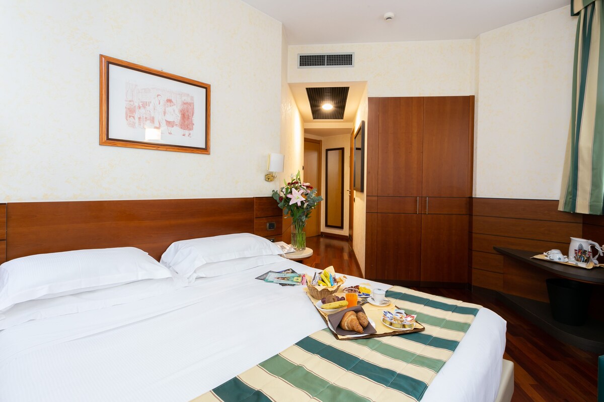 Een hotelkamer met een bed, een nachtkastje met een lamp, een lijst met foto aan de muur, en een houten garderobe.