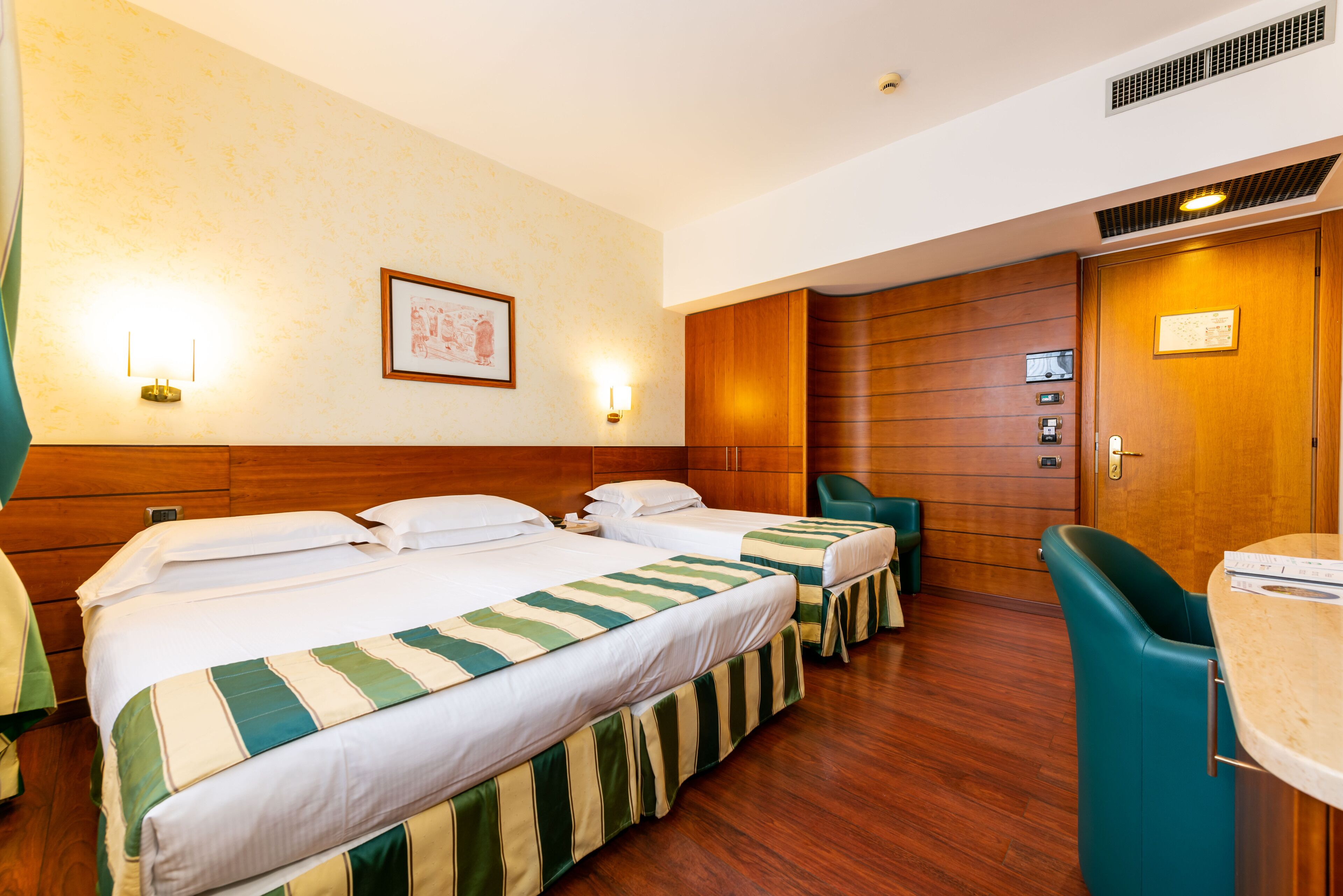 A hotel room with two beds, a wooden headboard, a wooden wardrobe, a green chair, and a small table.