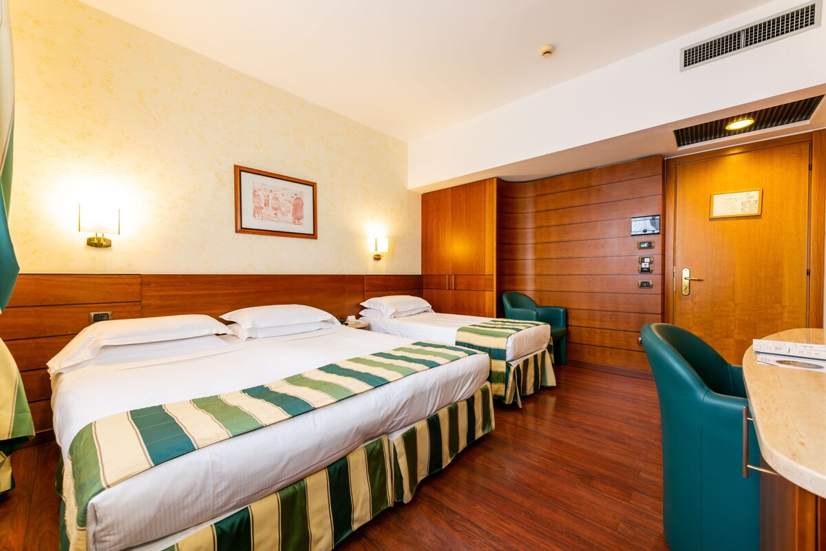 Een hotelkamer met twee bedden, een houten hoofdbord, een houten garderobe, een groene stoel en een kleine tafel.