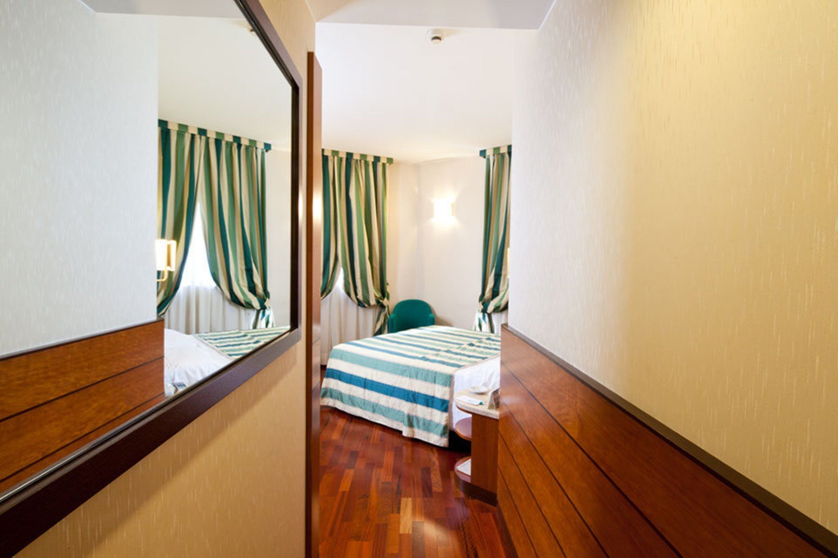 A narrow hotel room with a wooden floor, a bed with striped bedding, a green chair, and curtains with green and white stripes.