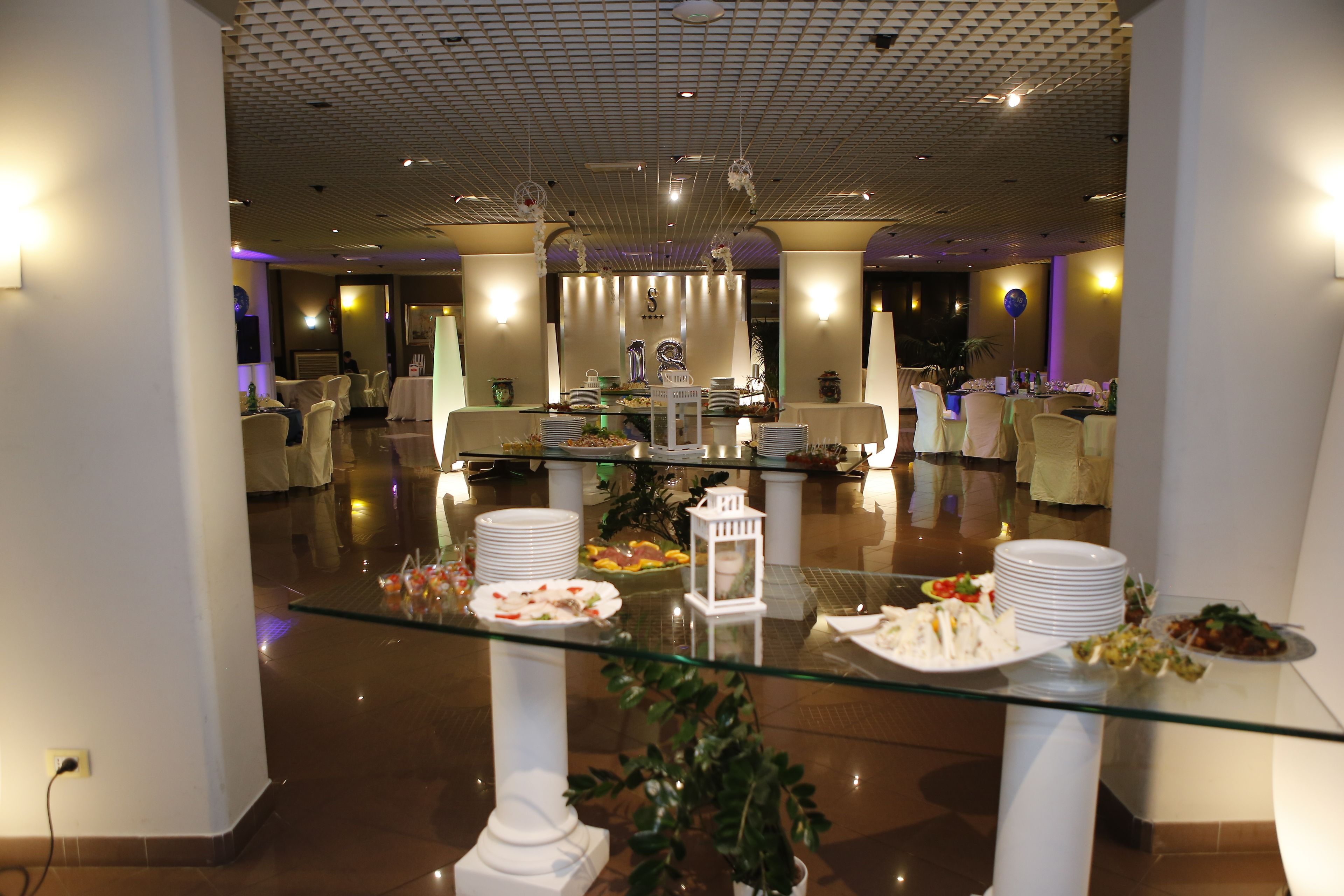 Un buffet est installé dans un hôtel, avec plusieurs tables, des chaises blanches et un présentoir en verre.