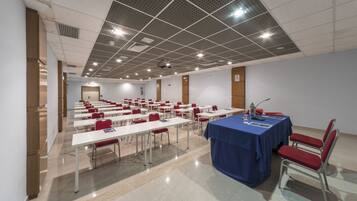 Una sala de conferencias con filas de mesas y sillas, un escenario con un atril, e iluminación empotrada en el techo.