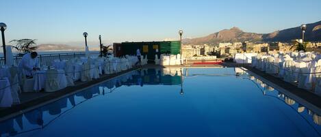 An outdoor pool with tables and chairs, a green building, and a mountainous backdrop.
