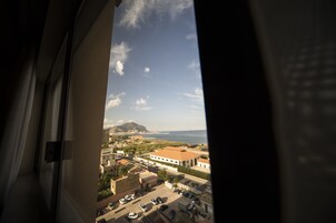 Une vue depuis une fenêtre donnant sur une ville côtière, avec ses bâtiments, ses voitures garées et une montagne à l’horizon.