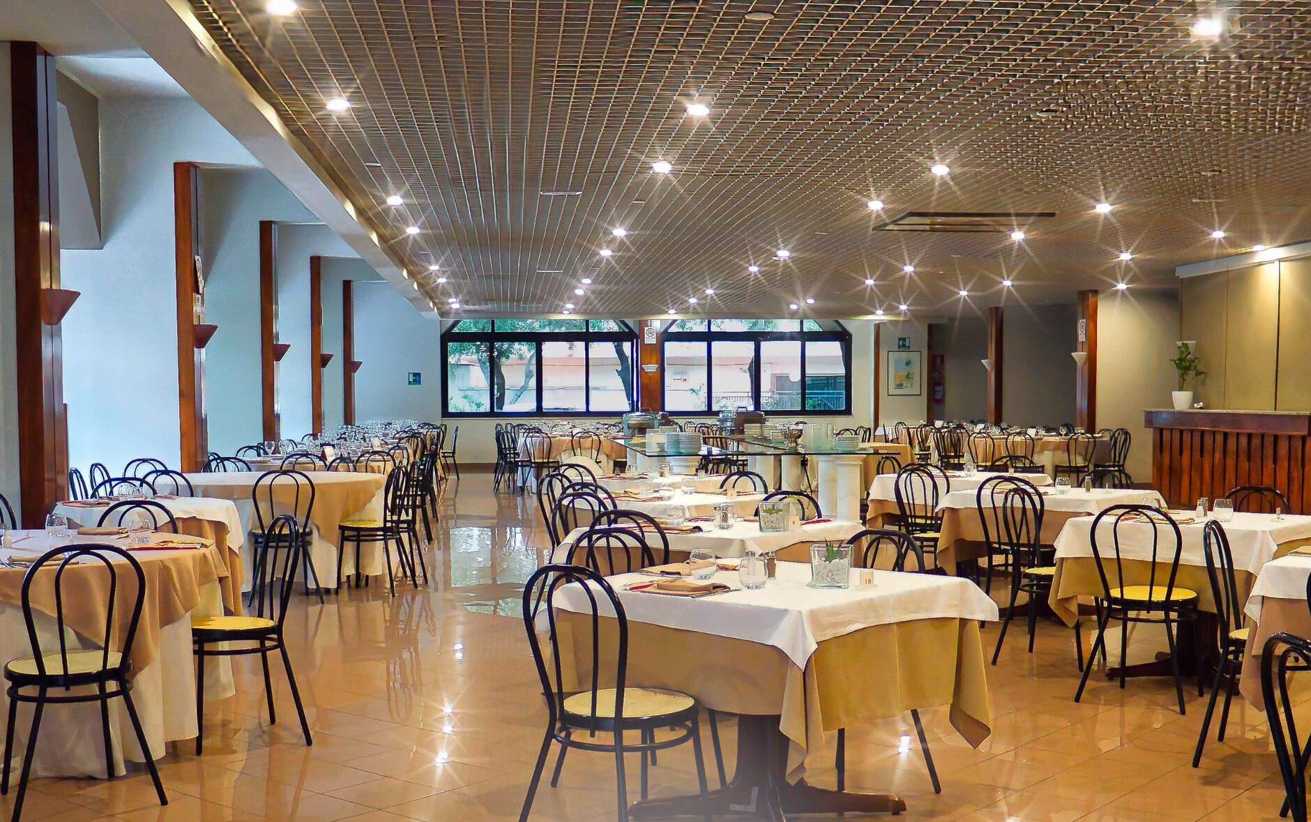 A spacious dining area with multiple tables and chairs, large windows, and a high ceiling with a grid pattern.