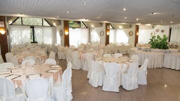 Une salle de banquet avec des tables rondes dressées pour le repas, des chaises blanches et une grande plante en pot.