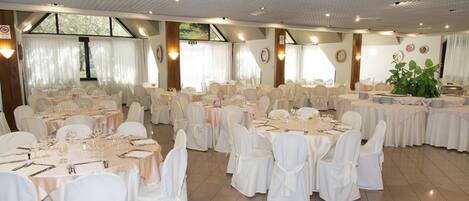 A banquet hall with round tables set for dining, white chairs, and a large potted plant.