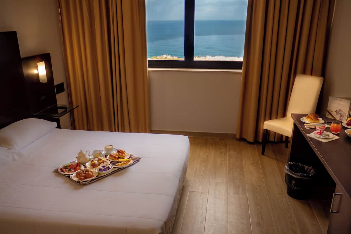 A hotel room with a bed, a dining table with breakfast, a chair, a desk, and a view of the ocean through a window.