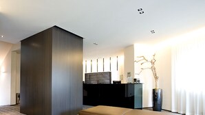 Un espace d’accueil moderne avec un comptoir d’accueil de couleur sombre, un coin salon avec un banc marron et un arbre décoratif dans un vase.