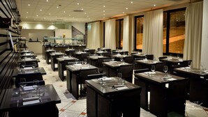 Un restaurant doté de nombreuses tables et chaises, d’un espace cuisine à l’arrière-plan et de grandes fenêtres avec des rideaux.