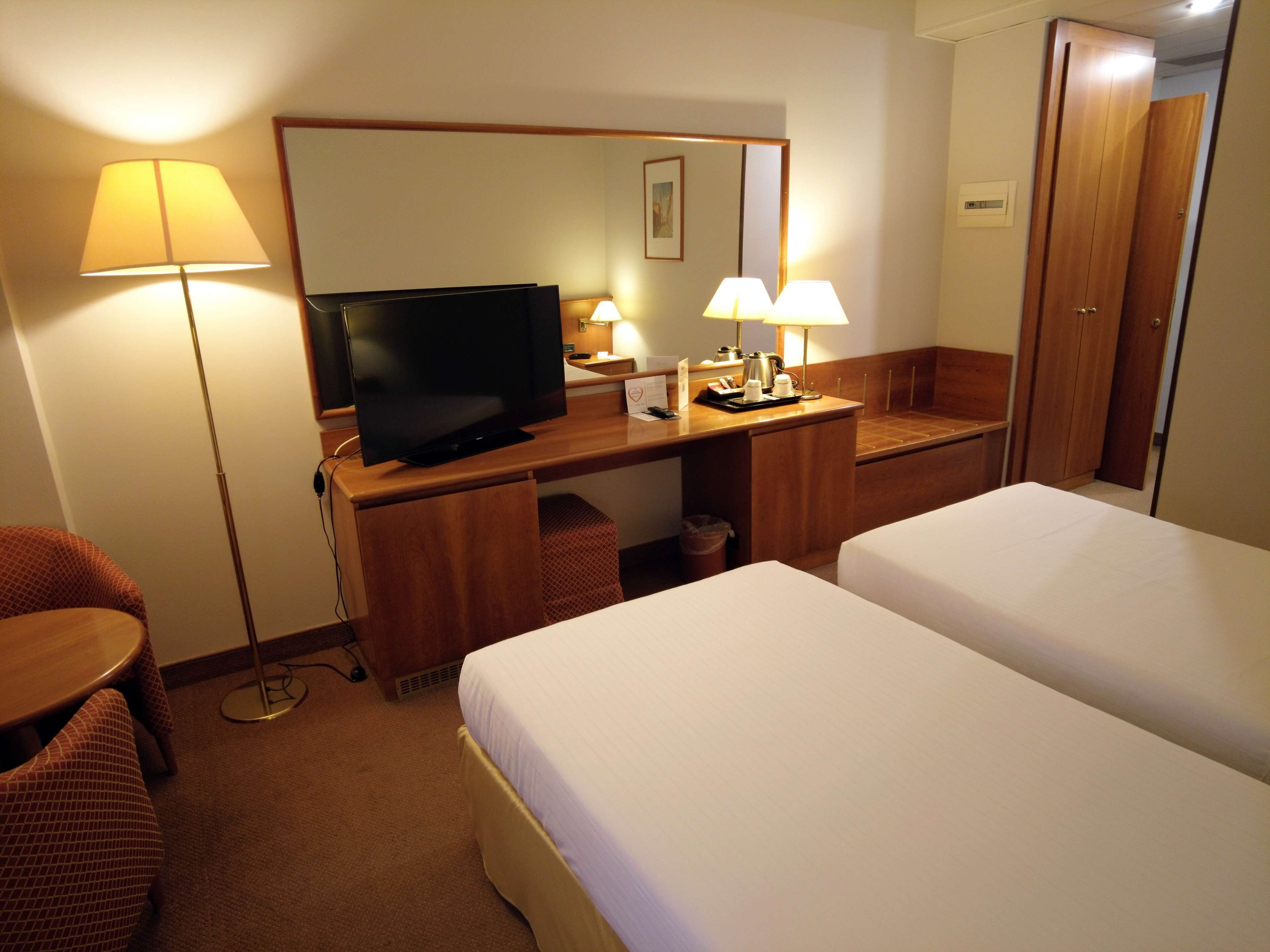 A hotel room with a TV, two beds, a desk with a lamp, a chair, and a trash bin.