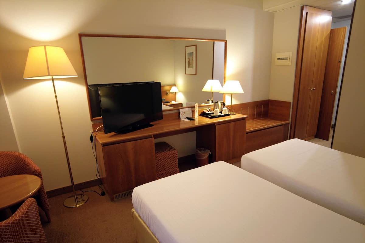 A hotel room with a TV, two beds, a desk with a lamp, a chair, and a trash bin.