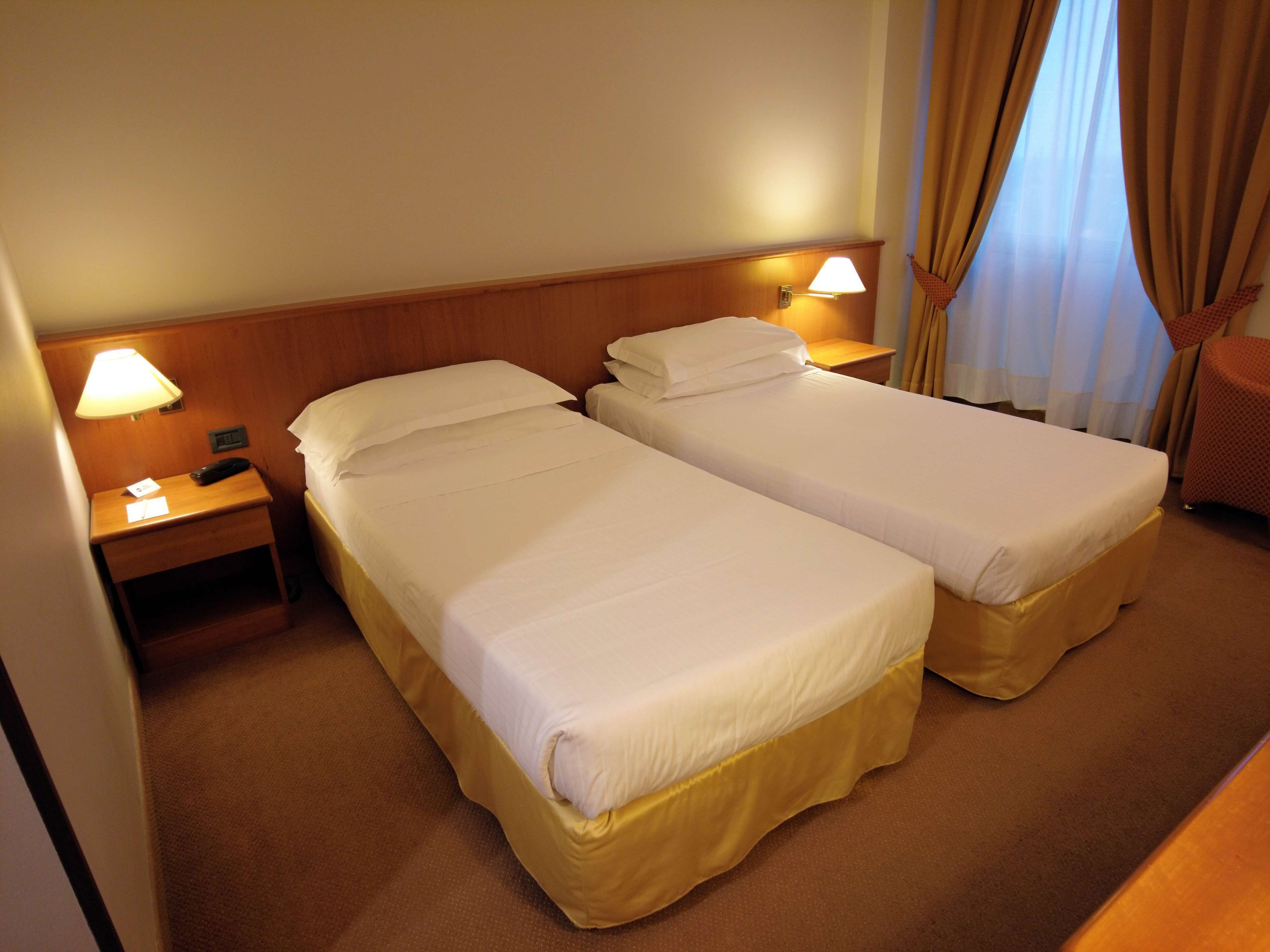 A hotel room with two single beds, a nightstand with a lamp, and a small table.