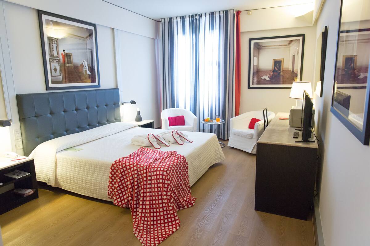 A hotel room with a large bed, a desk, a chair, a small table with a lamp, and a mirror.