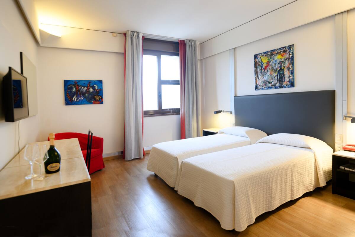 A hotel room with two beds, a desk with a bottle and glasses, a red chair, a window with curtains, and a painting on the wall.