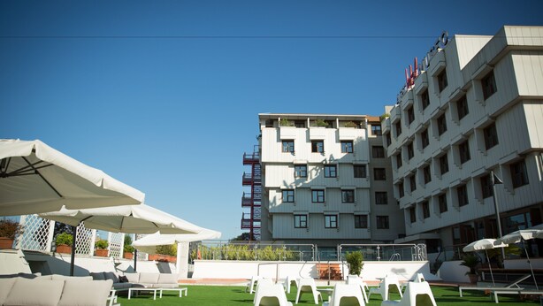 A rooftop terrace with white lounge chairs and umbrellas, a pool, and a building with balconies and flags. - Art Hotel Museo (Prato)