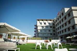 Terraza en la azotea con sillones blancos, sombrillas, piscina y un edificio con balcones y banderas.