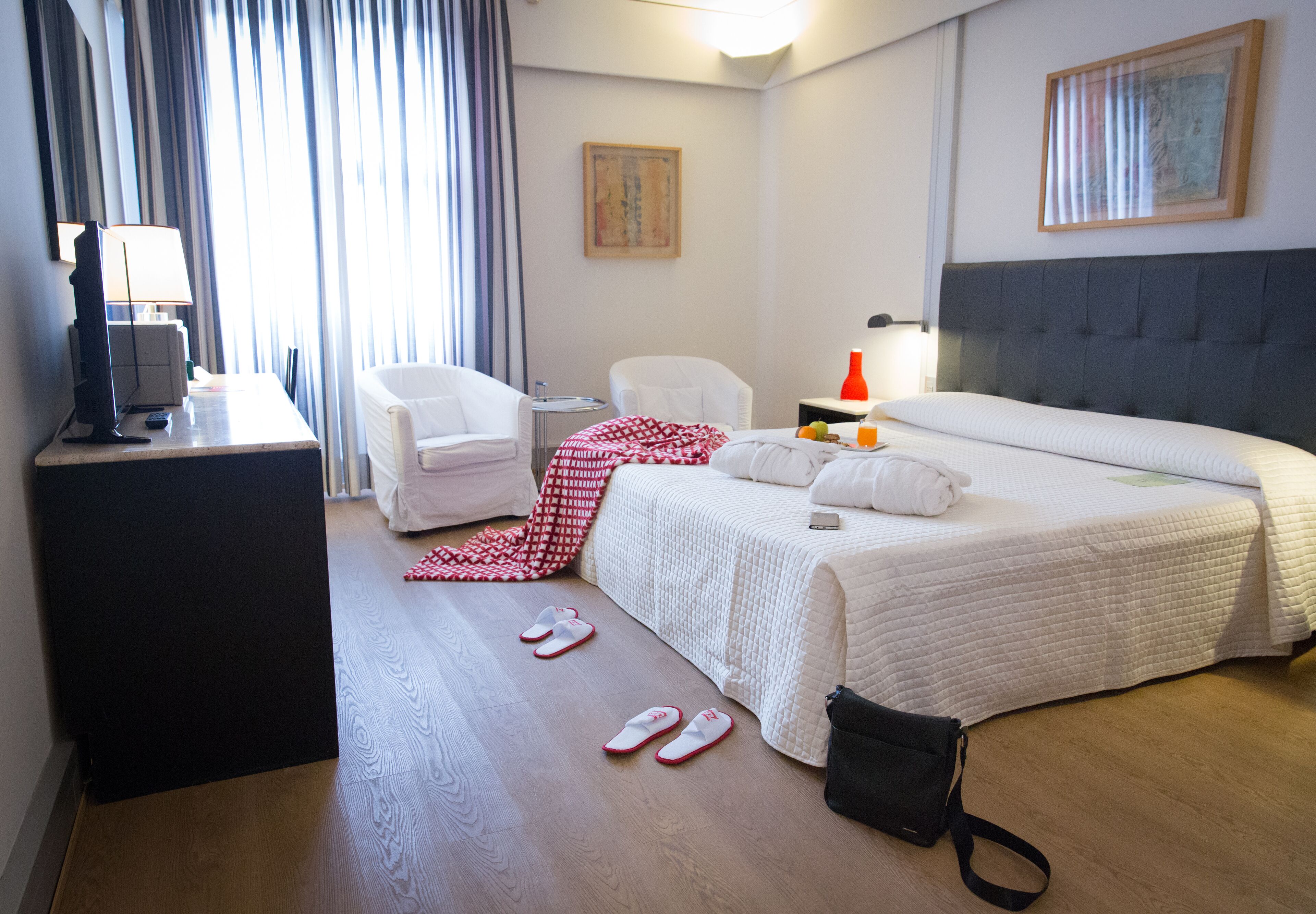 Una habitación de hotel con una cama, un escritorio con lámpara, una silla, una mesita de noche con lámpara y una ventana con cortinas.