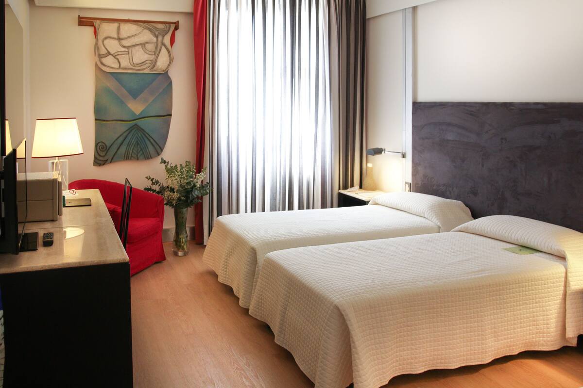 A hotel room with two beds, a desk with a lamp, a red chair, and a large abstract painting on the wall.
