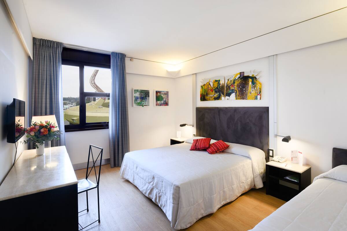 A hotel room with a bed, bedside tables, a desk, and a chair. There are paintings on the wall and a window with curtains.