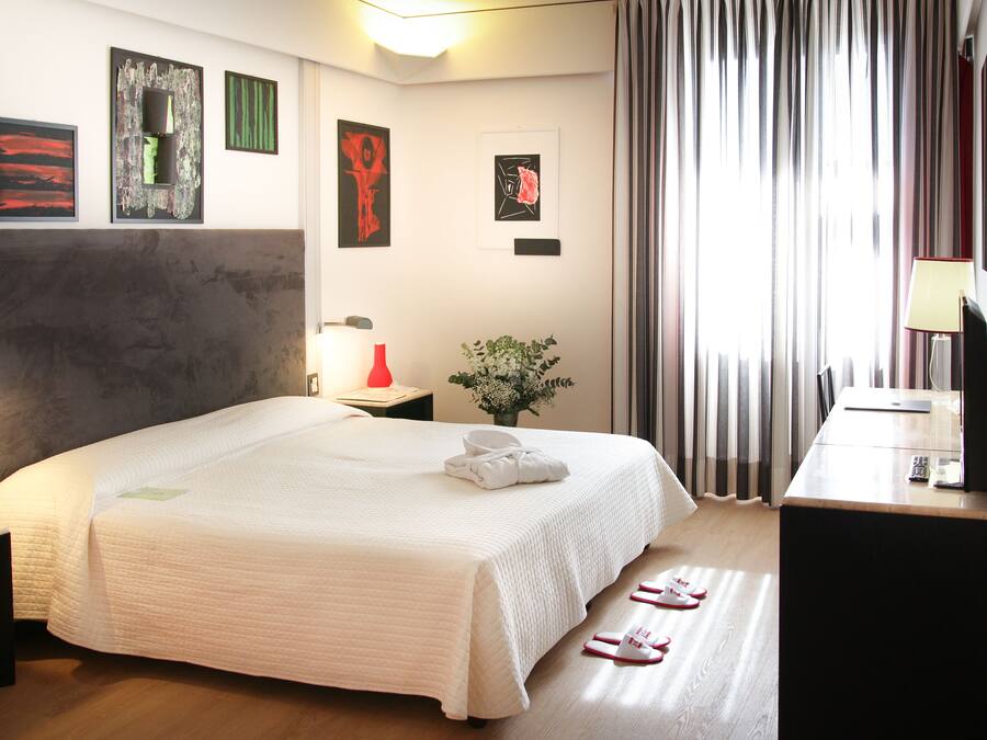 A hotel room with a large bed, a desk with a lamp, a TV, and artwork on the walls.