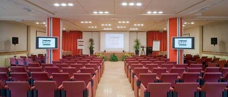 A conference room with rows of chairs, a podium, and a large screen at the front.