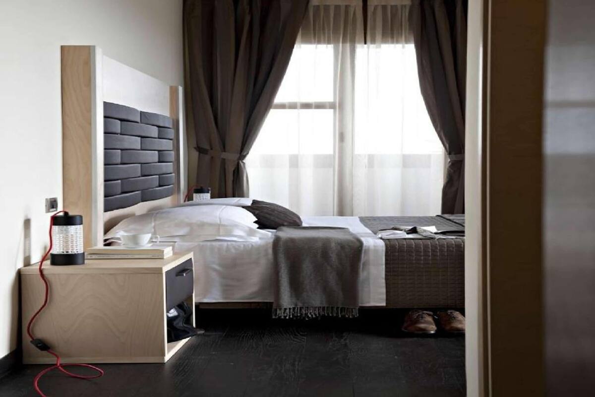 A modern hotel room with a dark wood floor, a bed with a grey headboard, a bedside table with a lamp, and a window with curtains.