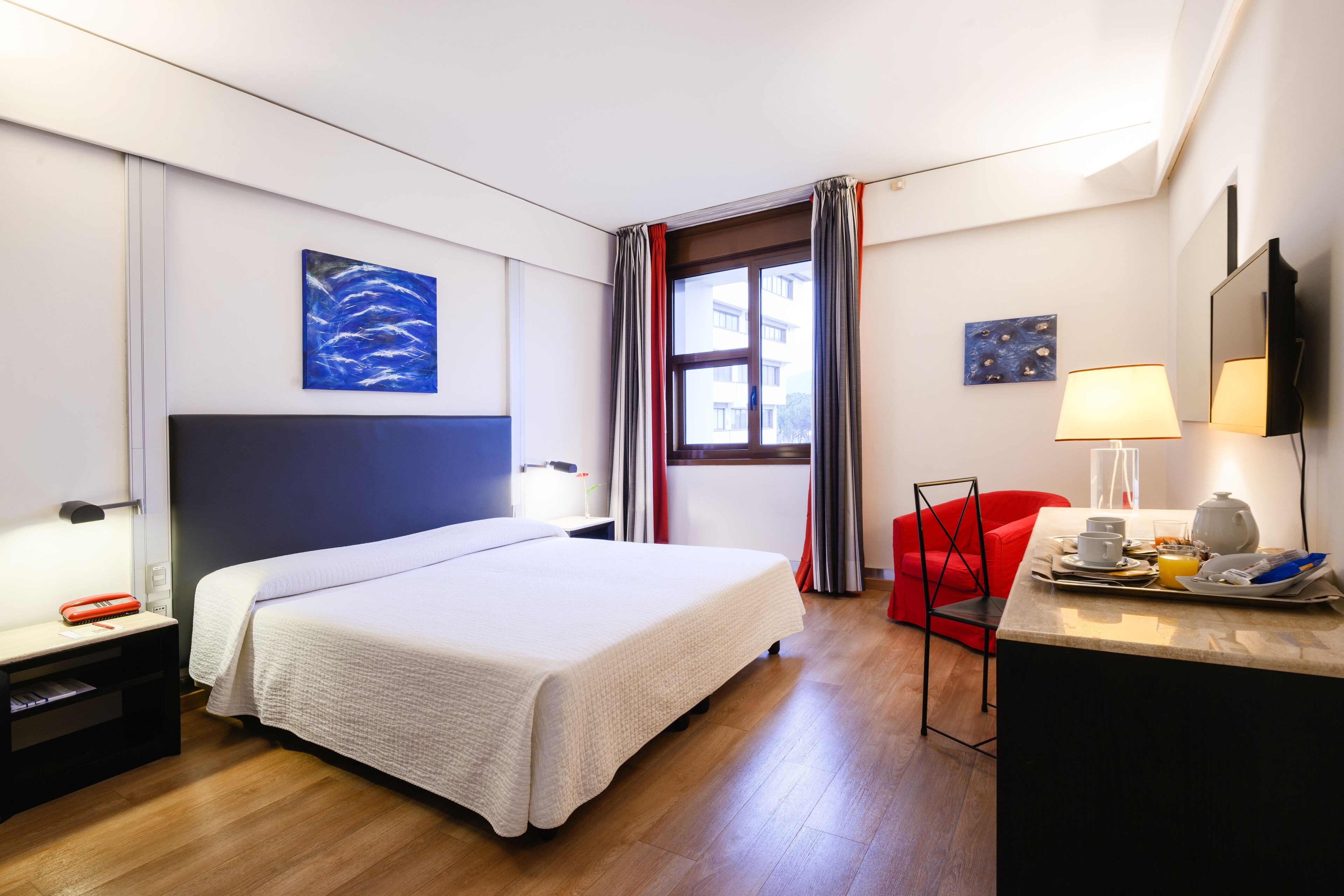 A hotel room with a bed, a desk with a lamp, a chair, and artwork on the walls.