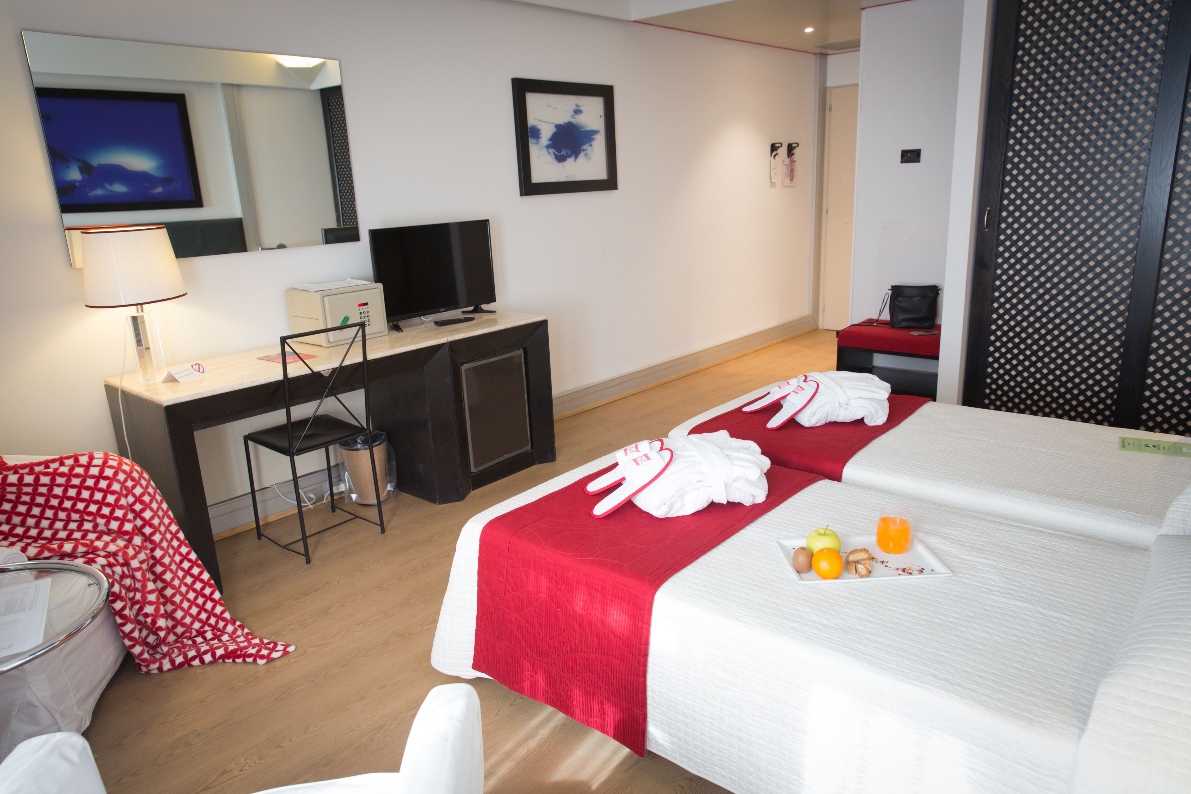 A hotel room with a bed, a desk, a chair, a TV, and a red and white checkered blanket.