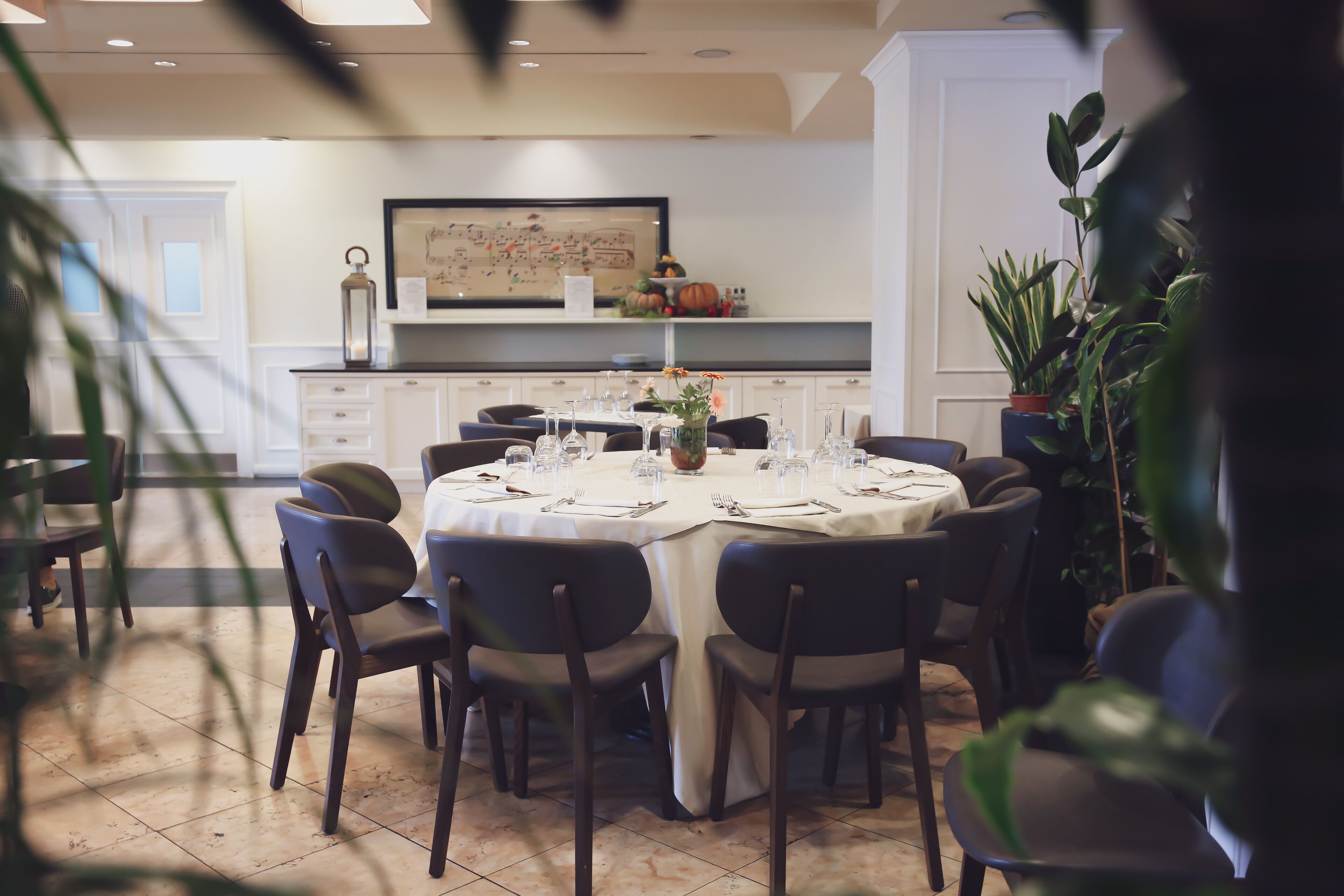 A dining area with a round table set for six, surrounded by chairs, in a room with white walls and a tiled floor.