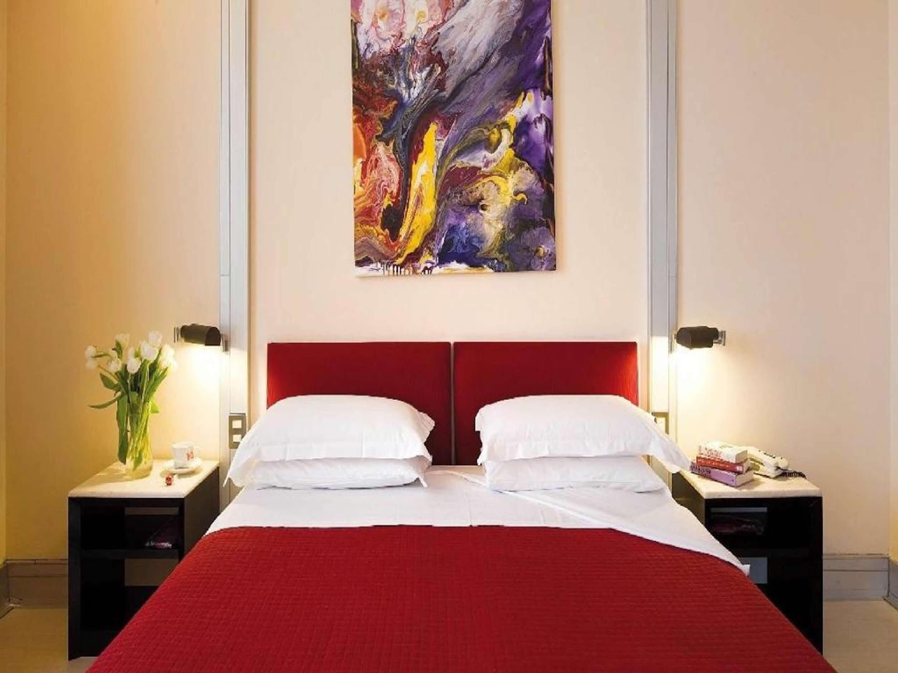 Une chambre d’hôtel avec un grand lit, une tête de lit rouge, des oreillers blancs et un tableau abstrait coloré au-dessus du lit. Il y a deux tables de chevet avec des lampes et un vase de fleurs sur l’une des tables.