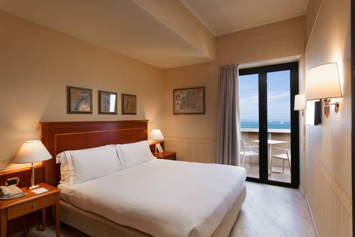 Une chambre d’hôtel avec un grand lit, deux tables de chevet avec des lampes, un téléphone et une fenêtre donnant sur la mer.