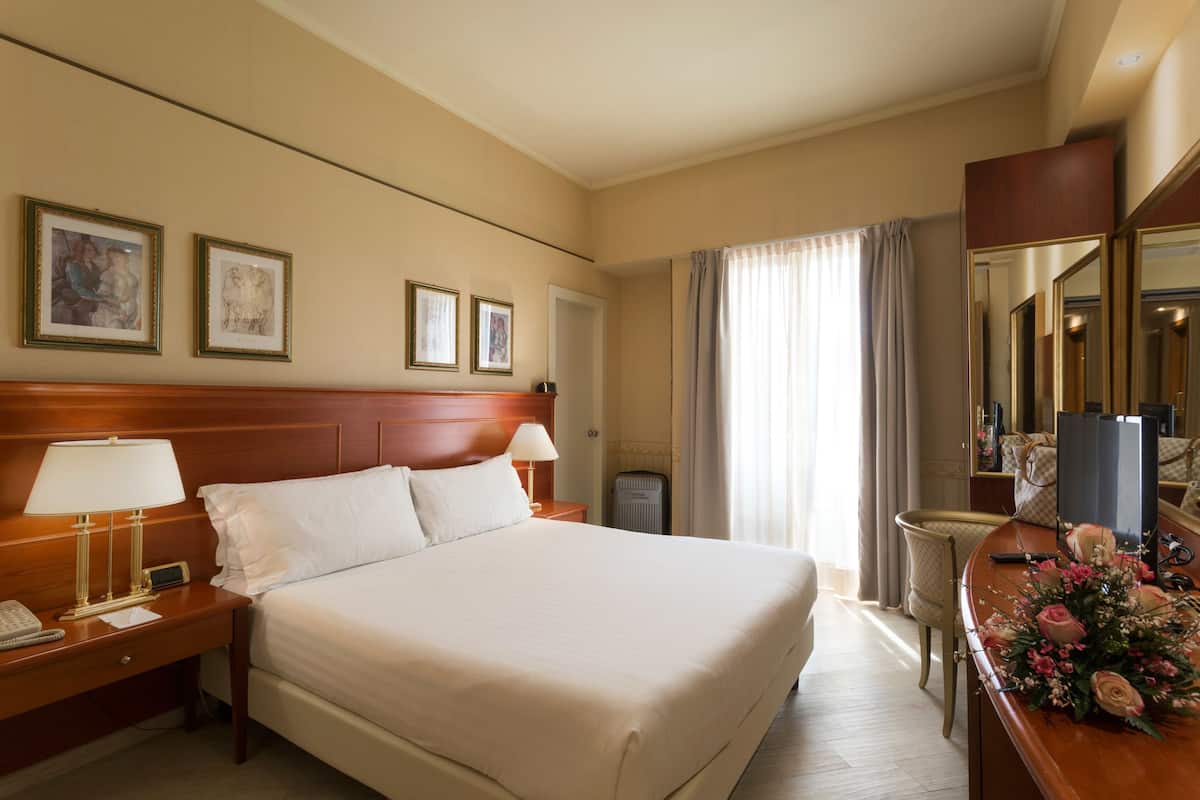 Une chambre d’hôtel avec un grand lit, des tables de chevet avec des lampes, un bureau avec un miroir et des tableaux encadrés au mur.